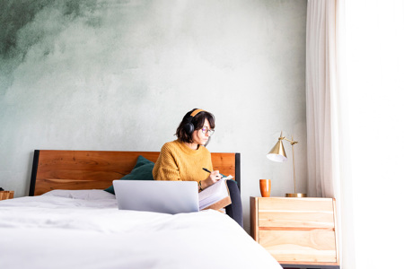 A woman sitting on her bed writing notes with her laptop open in front of her.