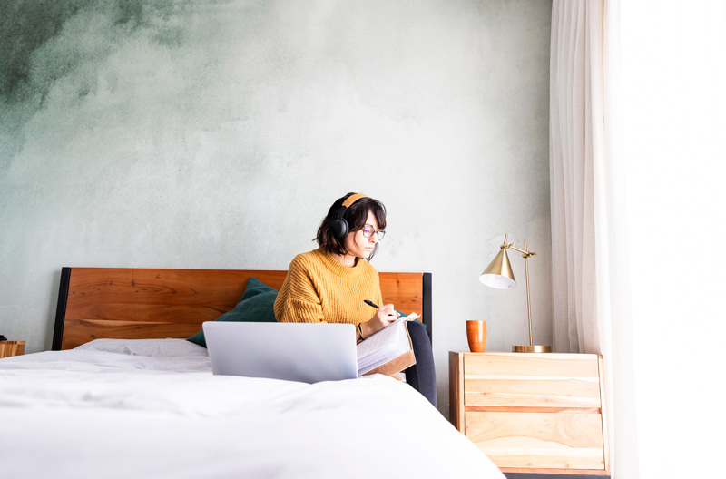 A woman sitting on her bed writing notes with her laptop open in front of her. 