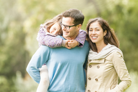 Two parents standing in a park with one of the parents holding their child on their back.