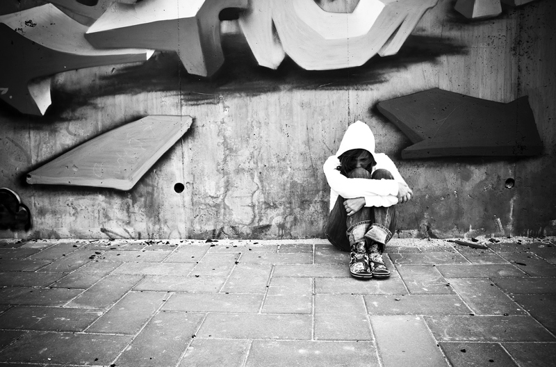 A woman sitting down leaning against a brick wall hugging her knees.