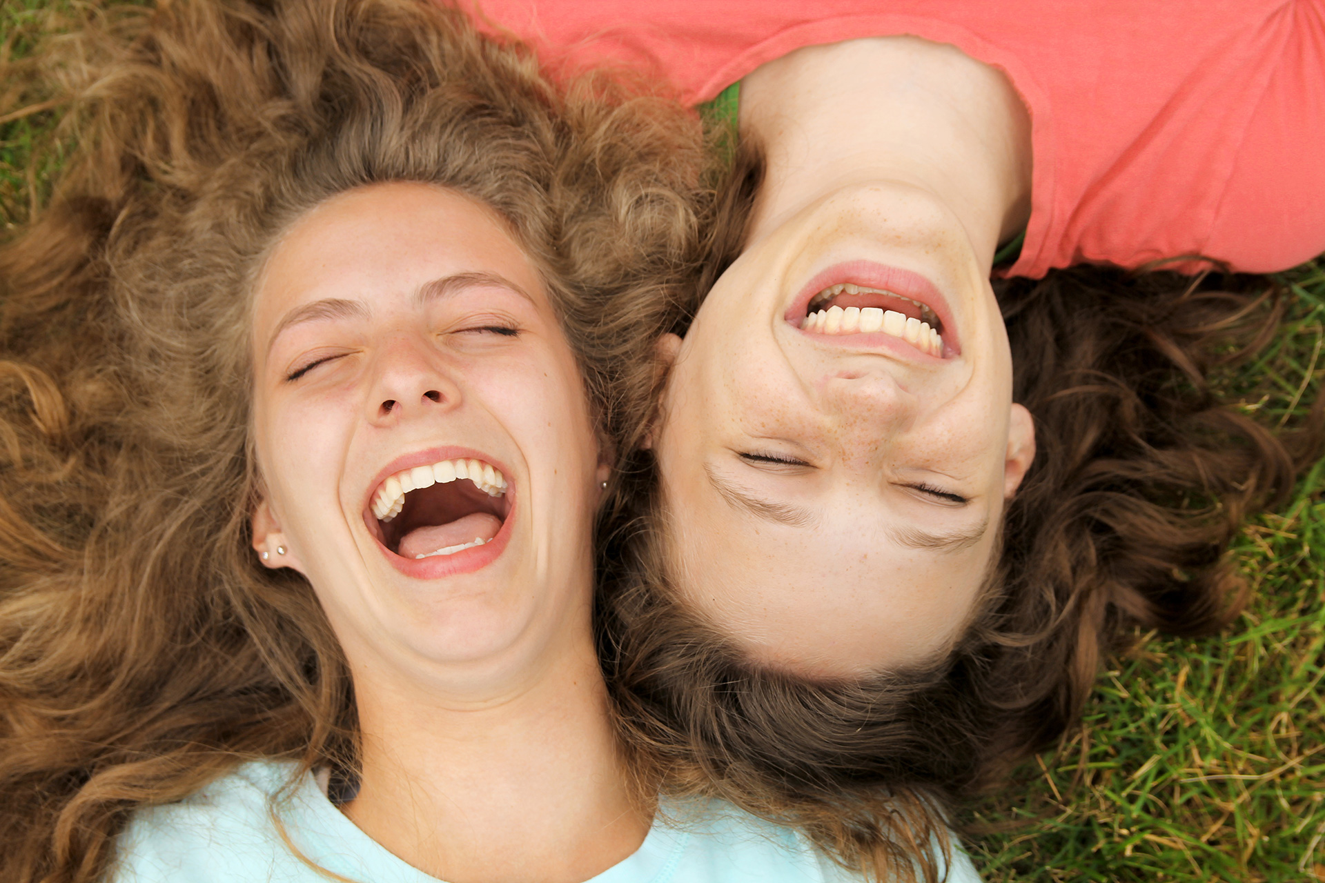 Two teenage girls laying down on the grass with their heads next to each other.