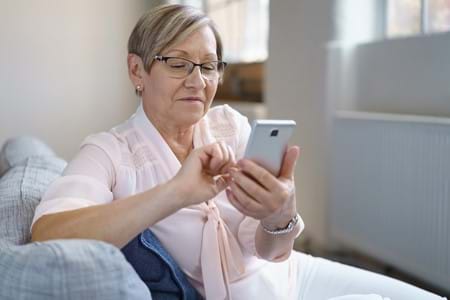 A woman wearing a light pink shirt sitting down on a couch scrolling on a phone.