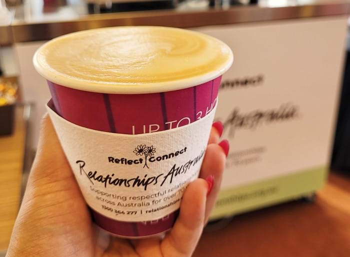 A takeaway coffee is held up in someone's hand in front of a coffee cart inside a building with a big glass window. The sleeve on the cup reads 'Reflect-Connect' and has the Relationships Australia logo
