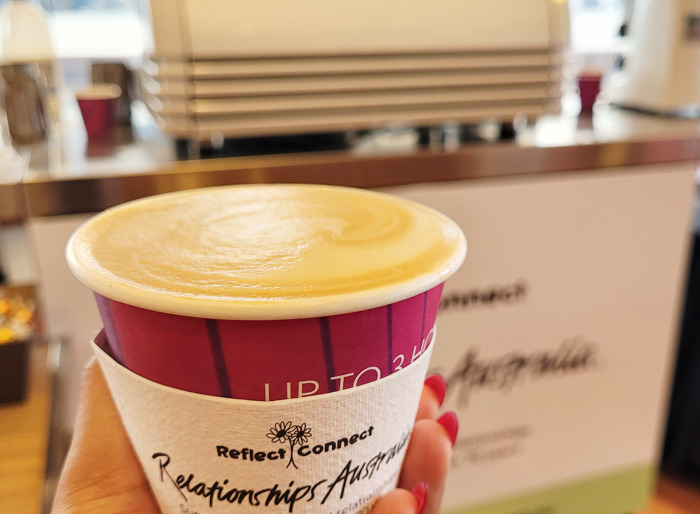 A takeaway coffee is held up in someone's hand in front of a coffee cart inside a building with a big glass window. The sleeve on the cup reads 'Reflect-Connect' and has the Relationships Australia logo