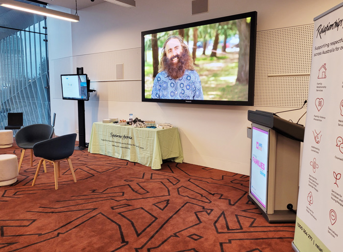 A conference room with chairs and low tables, a large video screen showing a smiling man standing in a park, a podium, pull-banner and a table covered with a green Relationships Australia tablecloth and a range of merchandise