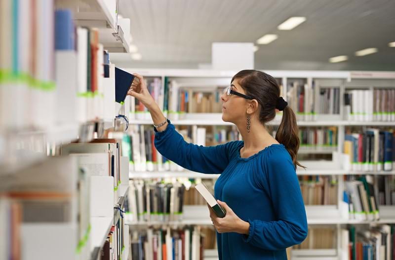 A woman in a blue shirt picking out books off a bookshelf in a library. 