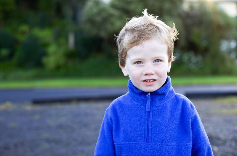 A young boy wearing a blue zip sweater standing in front of trees.