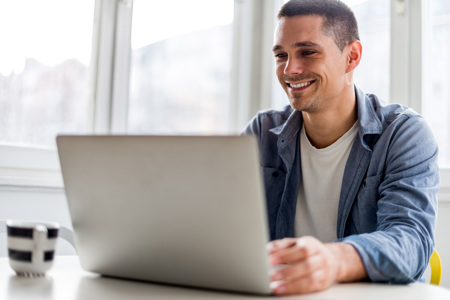 A man smiling as he sits at a table in front of an open laptop.