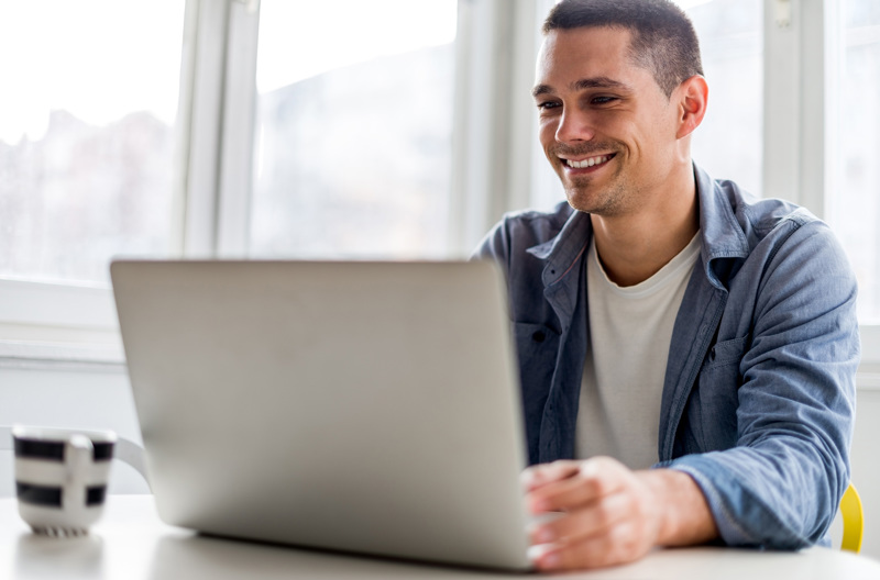 A man smiling as he sits at a table in front of an open laptop.