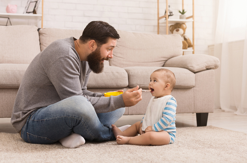 A young dad sitting on his living room carpet, feeding his infant child with a spoon.