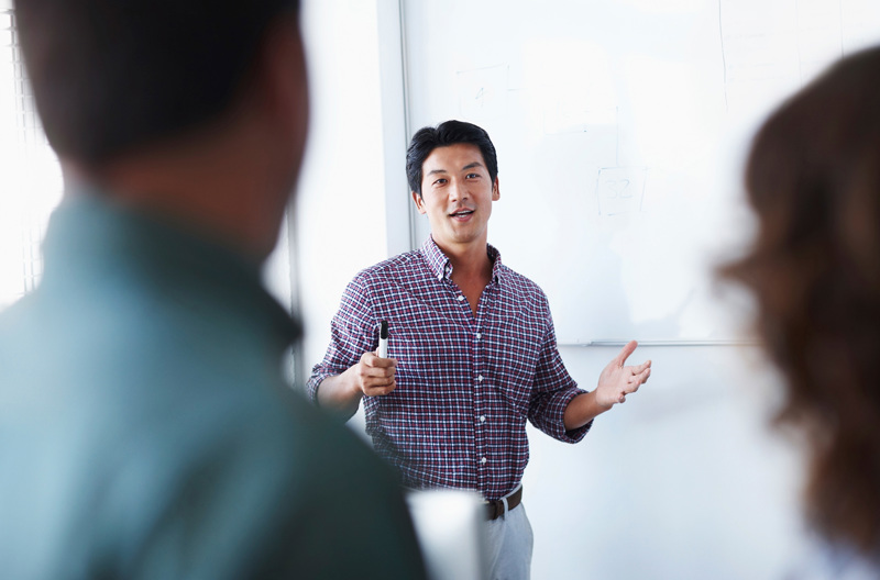 A teacher standing in front of a class holding a whiteboard marker.