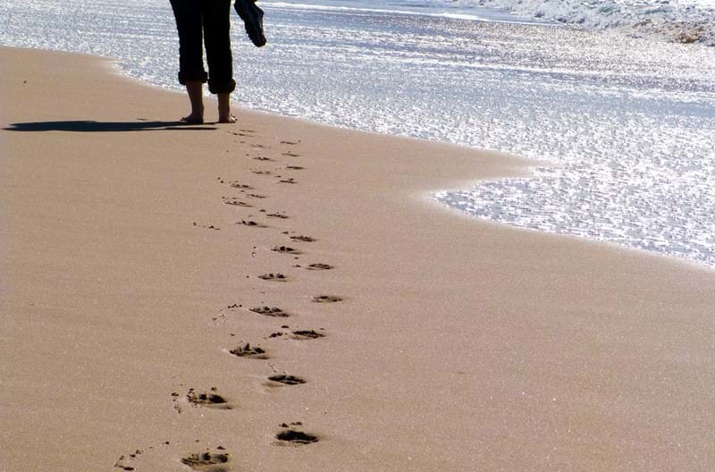 A person walking along a beach holding their shoes as they leave footmarks behind them.