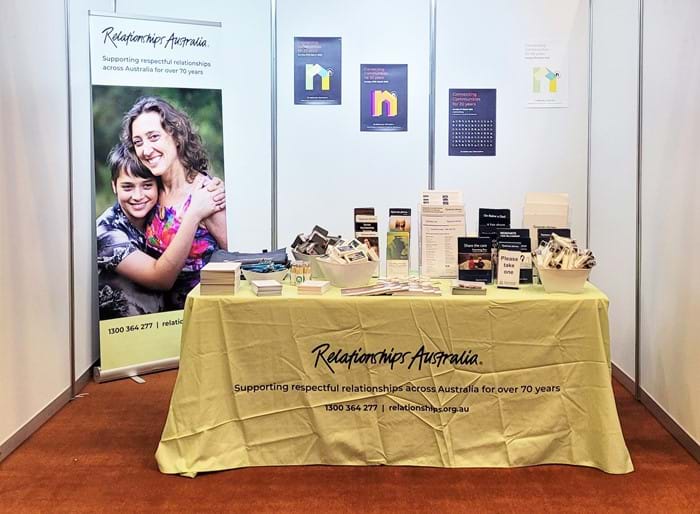 An indoor exhibition stand with a trestle table covered by a pale green Relationships Australia tablecloth, colourful posters on the back wall and a large pull-up banner with a picture of a mother and her teenage son. On the table are bowls and stands filled with brochures, flyers, and promotional merchandise
