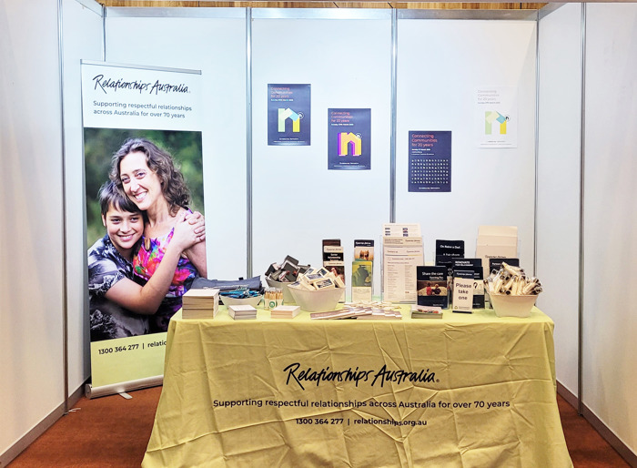 An indoor exhibition stand with a trestle table covered by a pale green Relationships Australia tablecloth, colourful posters on the back wall and a large pull-up banner with a picture of a mother and her teenage son. On the table are bowls and stands filled with brochures, flyers, and promotional merchandise