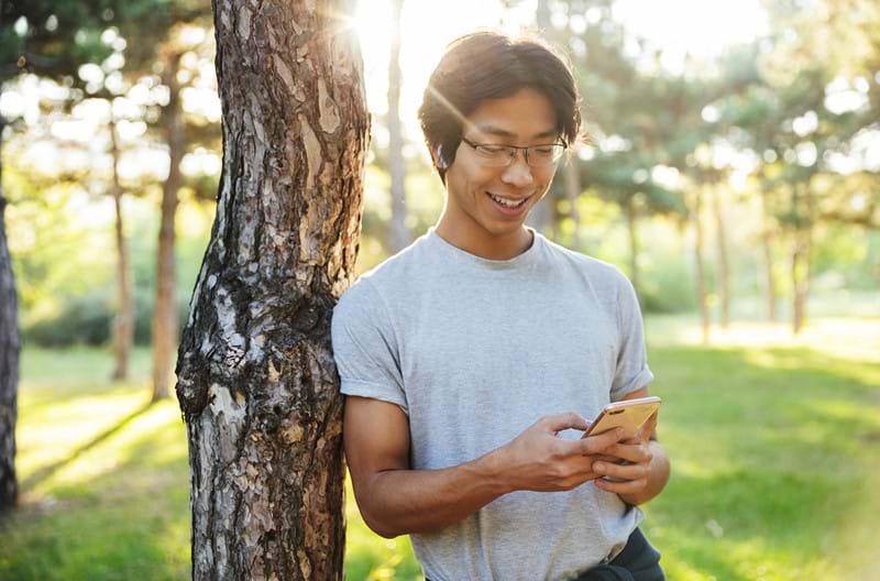 A man wearing a grey shirt leaning against a tree as he scrolls on his phone.