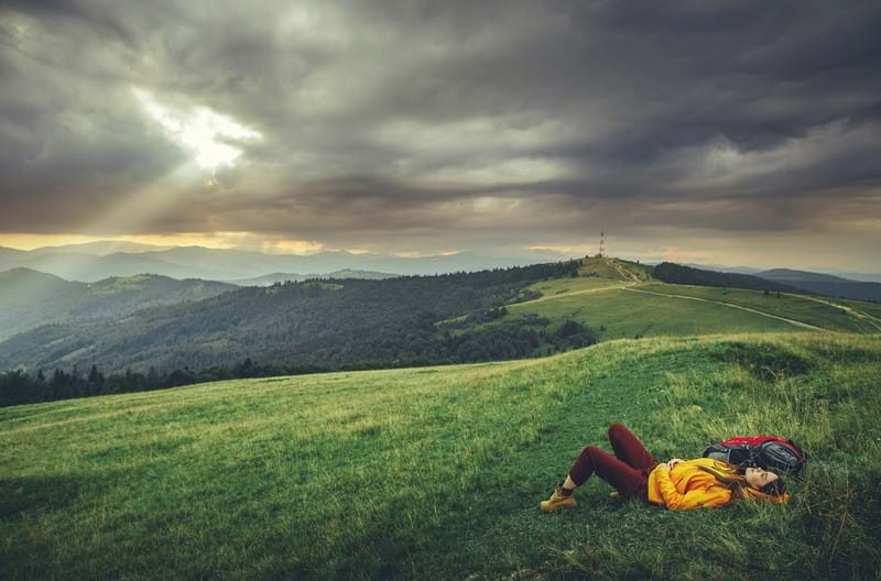 A person laying down wearing a yellow jacket on top of a grass hill looking up to a cloudy sky. 