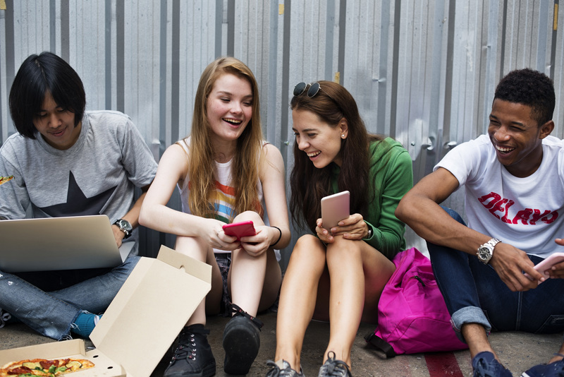 Four friends sitting on the ground eating pizza and playing on their phones.