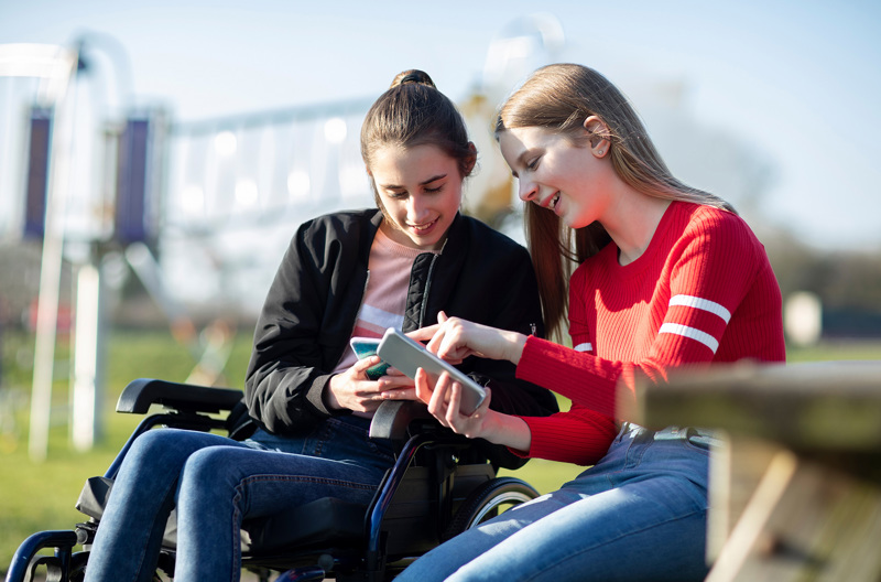 A girl sitting in a wheelchair next to a girl wearing a red sweater.