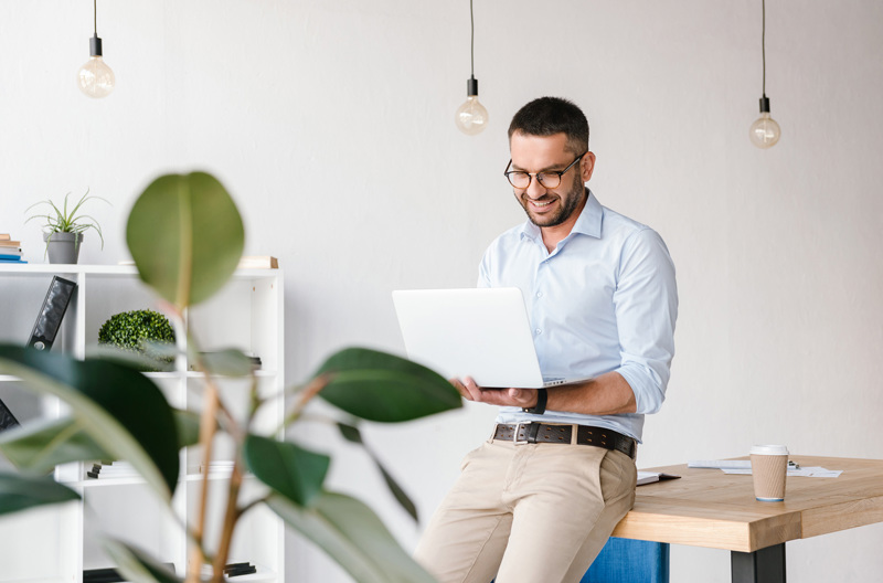 A man wearing a white business shirt leaning against a wooden table as he holds an open laptop.