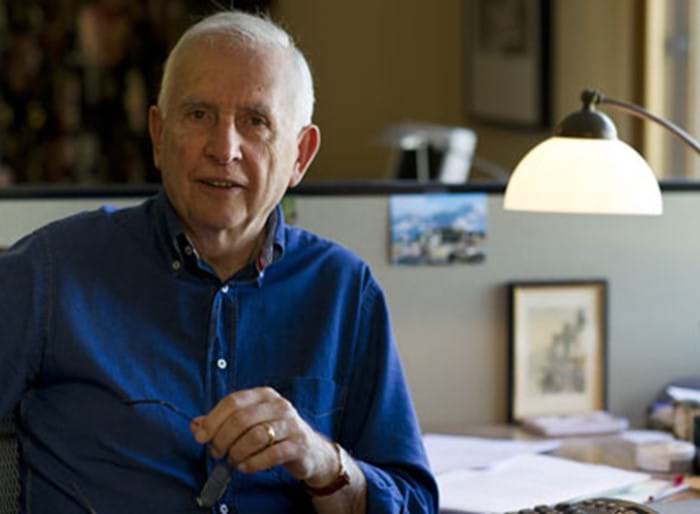 Hugh Mackay AO, a older Caucasian man with short white hair, smiling at the camera. He wears a dark blue collared shirt and is seated at a desk with a warm yellow lamp.