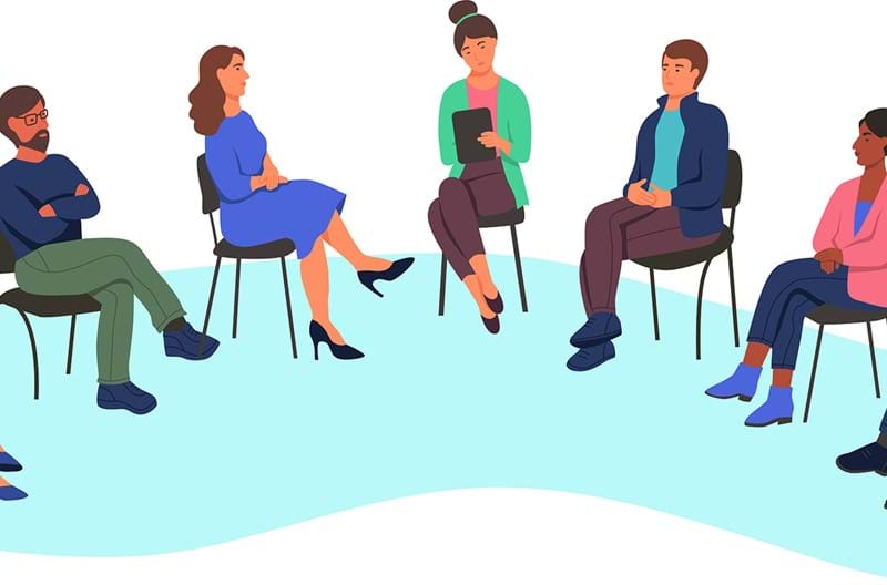 An illustration of a group of people sitting in a circle on chairs talking while one person takes notes on their clipboard. 
