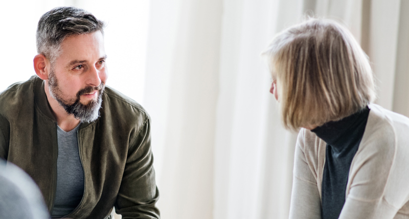 A man and a woman, seated indoors, having a friendly conversation. The man is Caucasian, with short, dark hair and a beard and wears a grey t-shirt under a khaki green jacket. The women is Caucasian with a shoulder length blond bob, and wears a dark, turtleneck top and a cream cardigan.