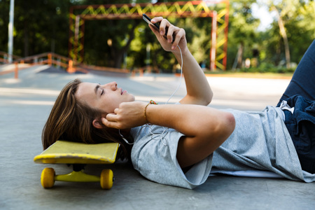 A teenager laying down on the ground resting his head on a yellow skateboard as he holds a phone in front of his face.