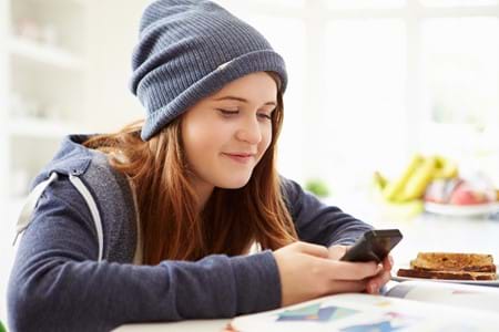 A teenage girl wearing a grey hoodie and beanie sitting down at a table smiling at a phone she's holding.